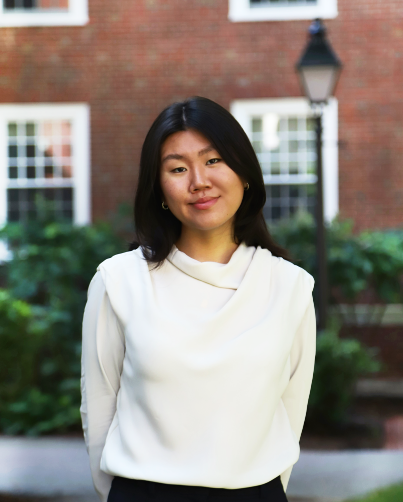 A person wearing a white blouse stands outdoors, smiling, with a brick building and greenery in the background.